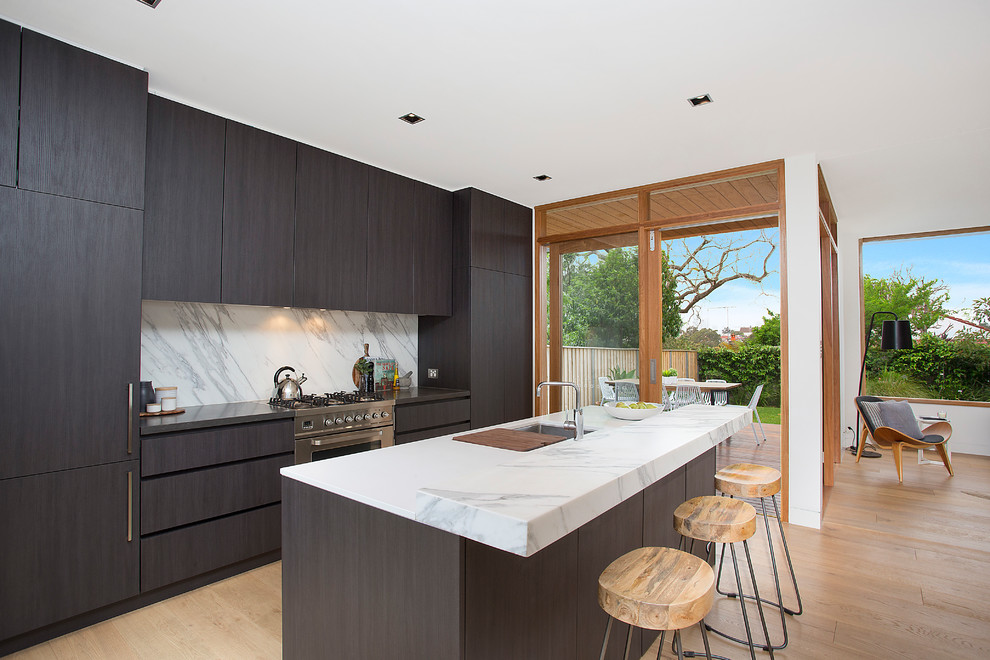 Inspiration for a beach style galley separate kitchen in Sydney with a double-bowl sink, flat-panel cabinets, black cabinets, stainless steel appliances, with island, grey splashback, light hardwood floors, beige floor and grey benchtop.