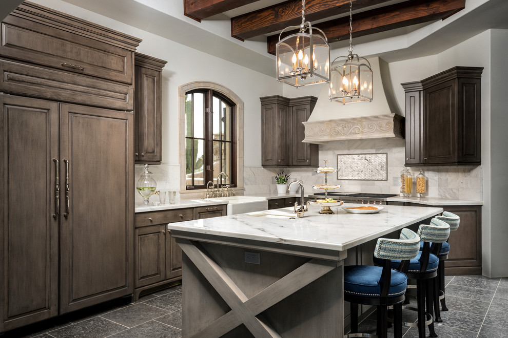 Design ideas for a large mediterranean l-shaped kitchen in Phoenix with a farmhouse sink, raised-panel cabinets, marble benchtops, stone tile splashback, limestone floors, with island, dark wood cabinets, beige splashback and panelled appliances.