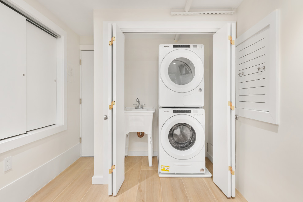 Design ideas for a small transitional single-wall laundry cupboard in New York with an utility sink, beige walls, light hardwood floors, a stacked washer and dryer and beige floor.