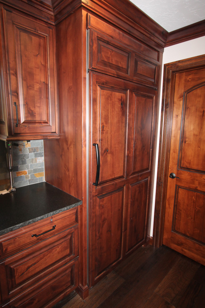 Large mountain style medium tone wood floor and brown floor kitchen photo with raised-panel cabinets, medium tone wood cabinets, granite countertops, multicolored backsplash, stone tile backsplash, paneled appliances, an island and black countertops