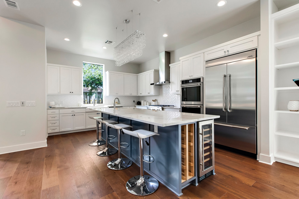 Large transitional l-shaped open plan kitchen in Austin with a farmhouse sink, shaker cabinets, white cabinets, quartzite benchtops, white splashback, stainless steel appliances, medium hardwood floors, with island, brown floor, white benchtop and glass sheet splashback.