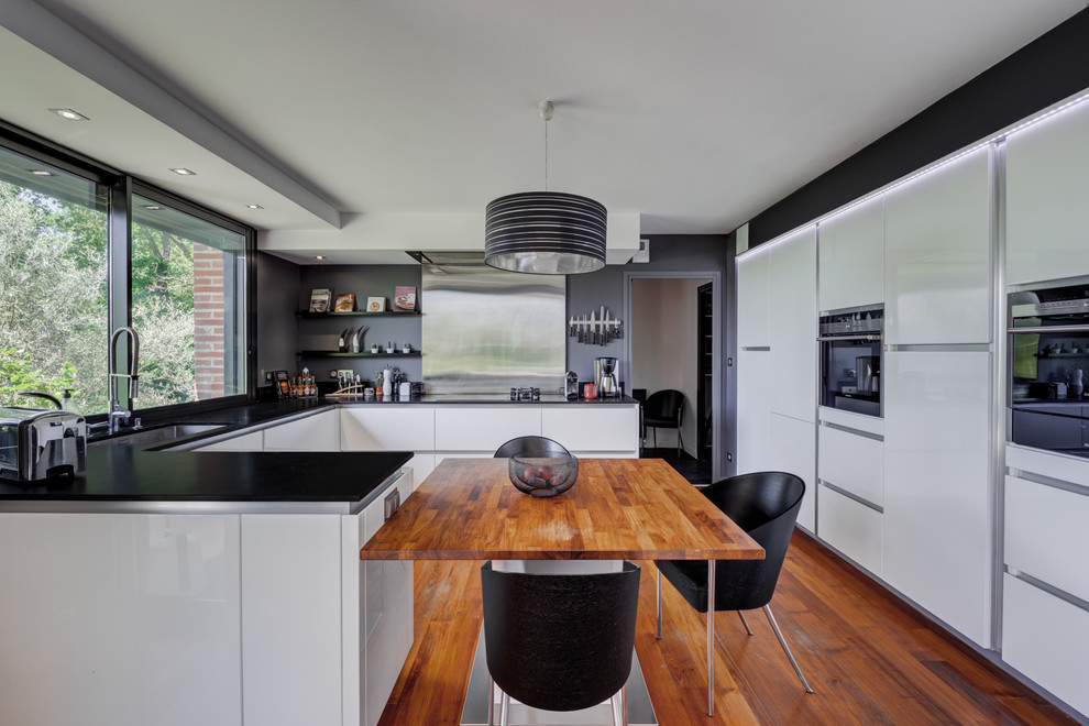 Design ideas for a contemporary u-shaped eat-in kitchen in Toulouse with an undermount sink, flat-panel cabinets, white cabinets, window splashback, medium hardwood floors, a peninsula and black benchtop.
