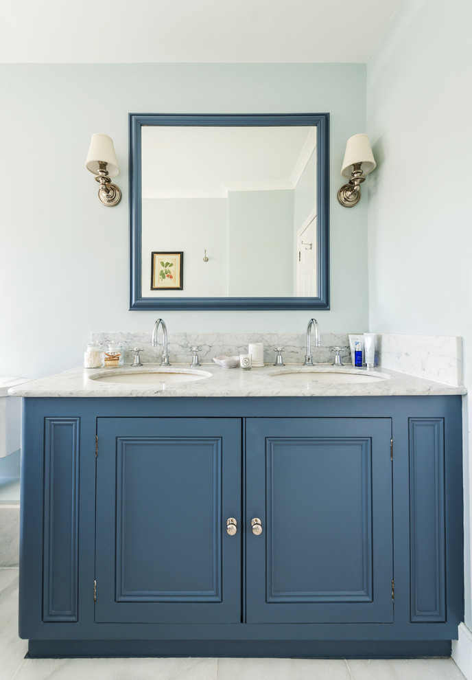 Photo of a mid-sized transitional master bathroom in London with recessed-panel cabinets, blue cabinets, a drop-in tub, a shower/bathtub combo, a two-piece toilet, white tile, stone tile, blue walls, marble floors, an undermount sink and marble benchtops.