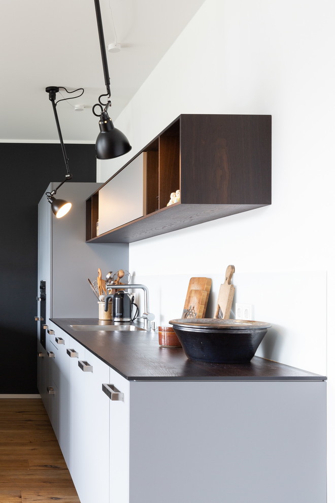 Mid-sized contemporary galley eat-in kitchen in Cologne with a single-bowl sink, flat-panel cabinets, grey cabinets, wood benchtops, white splashback, glass tile splashback, black appliances, dark hardwood floors, with island, brown floor and brown benchtop.