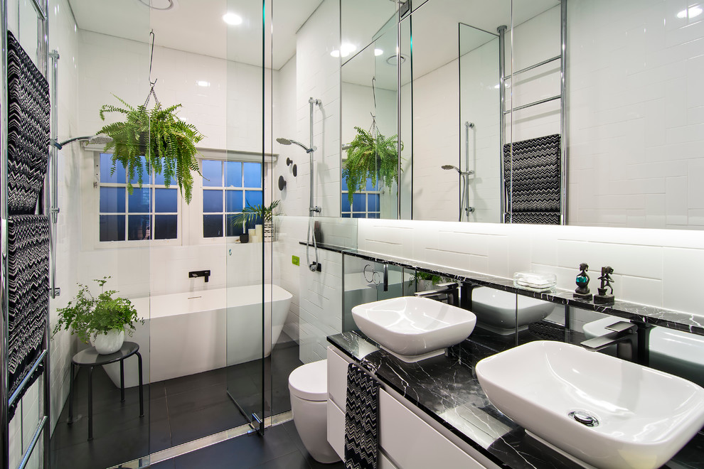 This is an example of a small contemporary master wet room bathroom in Sydney with white cabinets, white tile, white walls, a vessel sink, a hinged shower door, a freestanding tub, a one-piece toilet, subway tile, ceramic floors, marble benchtops, black floor, black benchtops and flat-panel cabinets.