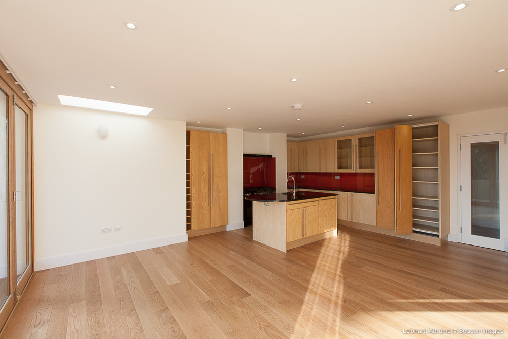 This is an example of a large eclectic l-shaped open plan kitchen in London with an integrated sink, flat-panel cabinets, light wood cabinets, granite benchtops, red splashback, glass sheet splashback, black appliances, light hardwood floors and with island.