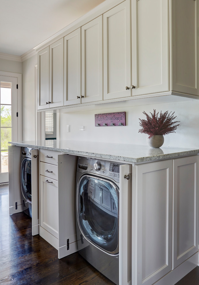 Inspiration for a mid-sized traditional u-shaped utility room in Chicago with a farmhouse sink, shaker cabinets, white cabinets, granite benchtops, white walls, dark hardwood floors, a concealed washer and dryer, brown floor and multi-coloured benchtop.