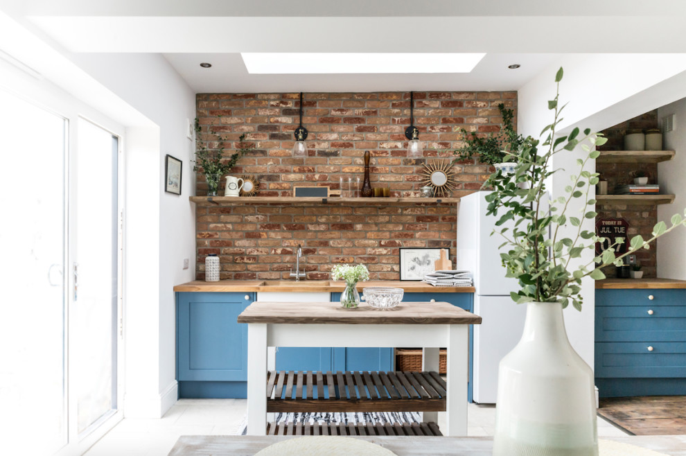 Photo of a mid-sized industrial single-wall eat-in kitchen with a farmhouse sink, shaker cabinets, blue cabinets, wood benchtops, red splashback, brick splashback, stainless steel appliances, cement tiles, with island, grey floor and brown benchtop.