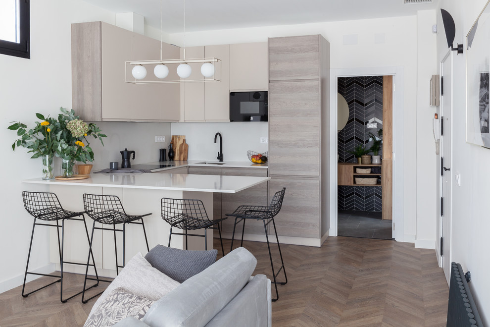 This is an example of a mid-sized scandinavian u-shaped open plan kitchen in Madrid with an undermount sink, flat-panel cabinets, beige cabinets, quartz benchtops, beige splashback, limestone splashback, black appliances, laminate floors, a peninsula, brown floor and beige benchtop.