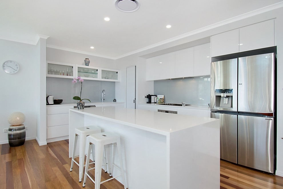 This is an example of a beach style open plan kitchen in Other with an undermount sink, white cabinets, quartz benchtops, grey splashback, glass sheet splashback, stainless steel appliances, medium hardwood floors, with island and white benchtop.