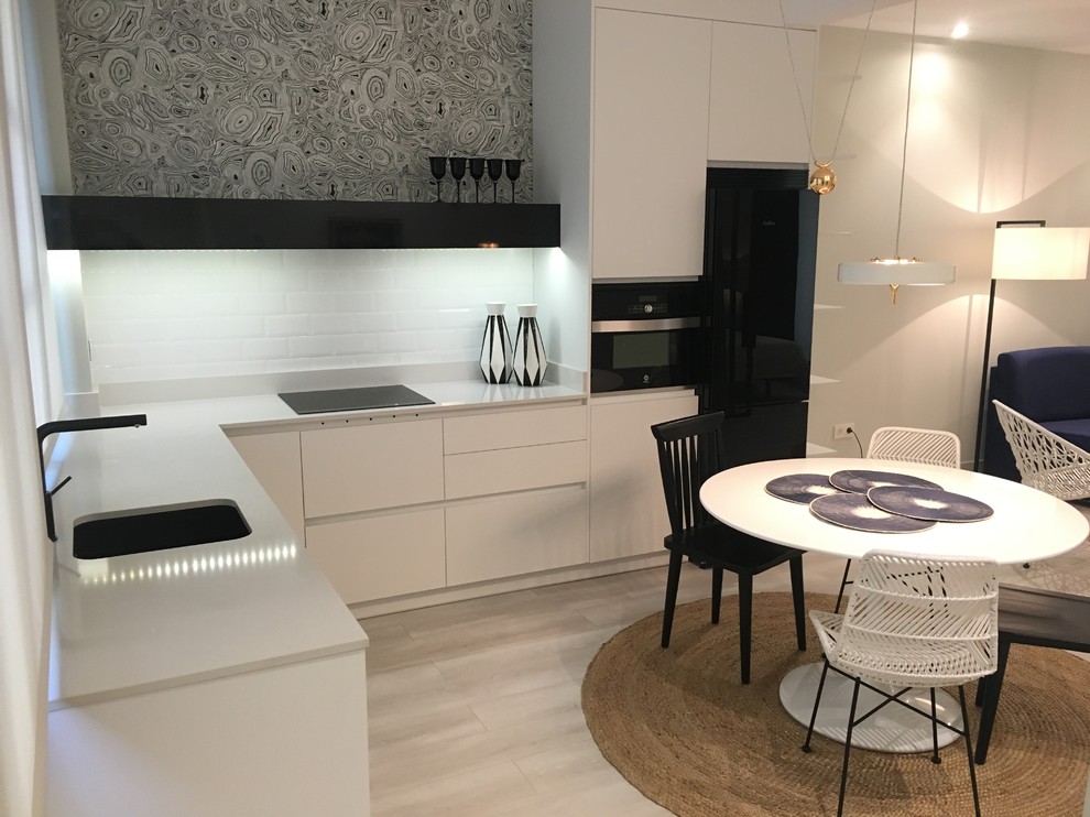 This is an example of a mid-sized modern l-shaped open plan kitchen in Madrid with an undermount sink, flat-panel cabinets, white cabinets, quartz benchtops, black splashback, subway tile splashback, black appliances and light hardwood floors.