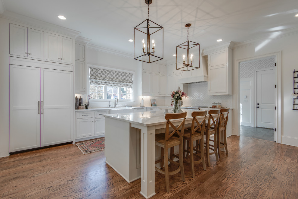 Design ideas for a large transitional l-shaped kitchen in Nashville with a farmhouse sink, recessed-panel cabinets, white cabinets, quartz benchtops, white splashback, porcelain splashback, panelled appliances, medium hardwood floors, with island and brown floor.