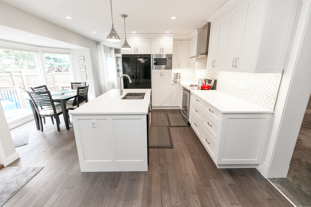 Example of a large transitional l-shaped medium tone wood floor and brown floor eat-in kitchen design in Calgary with an undermount sink, shaker cabinets, white cabinets, quartz countertops, white backsplash, an island and black appliances