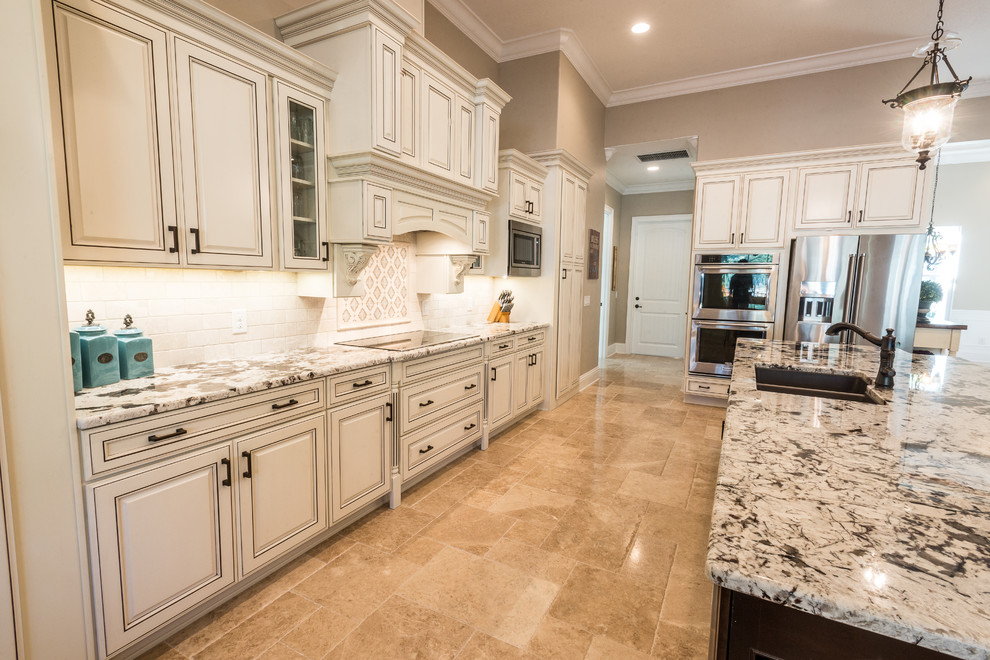 Photo of a large traditional l-shaped kitchen in Orlando with an undermount sink, raised-panel cabinets, white cabinets, granite benchtops, white splashback, subway tile splashback, stainless steel appliances, travertine floors and with island.