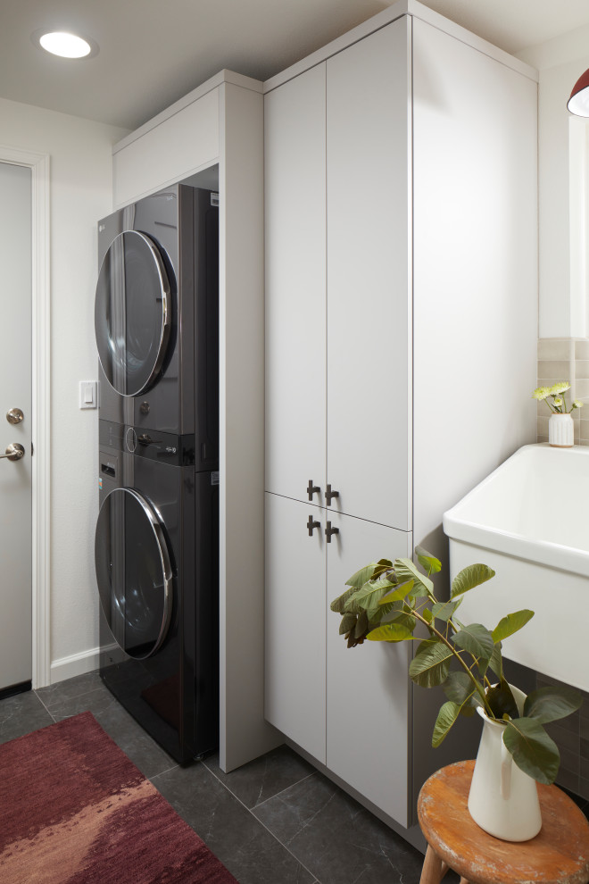 Inspiration for a small transitional dedicated laundry room in Seattle with an utility sink, flat-panel cabinets, grey cabinets, beige splashback, ceramic splashback, white walls, porcelain floors, a stacked washer and dryer and black floor.