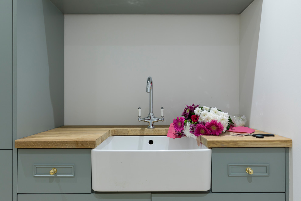 Photo of a small traditional single-wall dedicated laundry room in London with a farmhouse sink, shaker cabinets, green cabinets, wood benchtops, grey walls, slate floors, beige floor and brown benchtop.