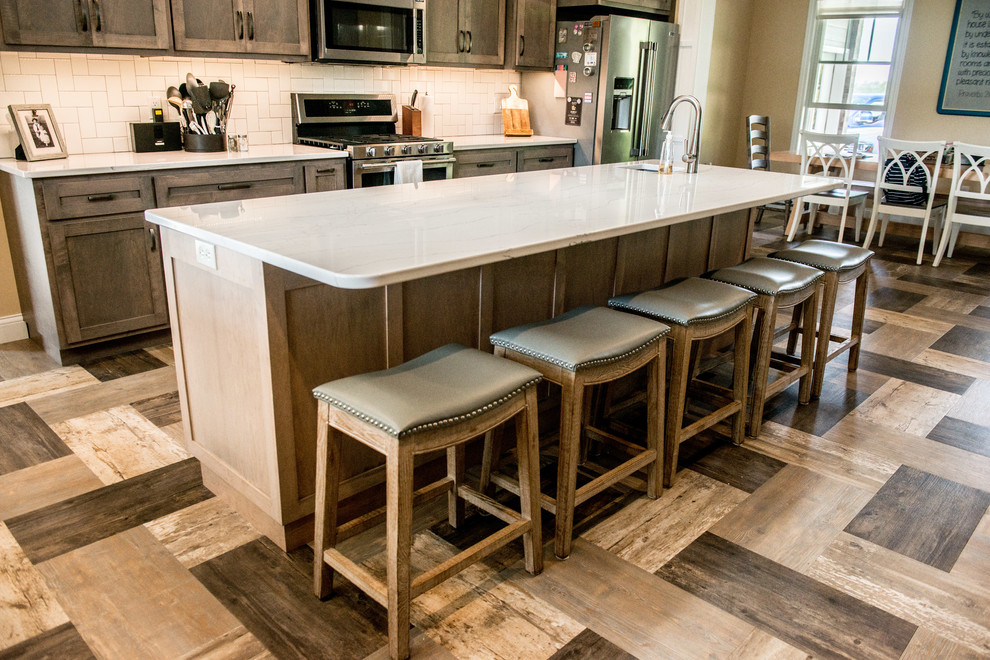 Example of a large transitional l-shaped vinyl floor and multicolored floor open concept kitchen design with a farmhouse sink, shaker cabinets, gray cabinets, quartz countertops, white backsplash, subway tile backsplash, stainless steel appliances, an island and white countertops