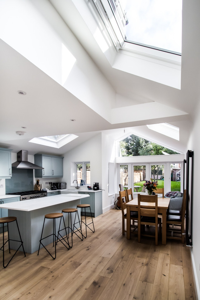 This is an example of a mid-sized contemporary l-shaped open plan kitchen in Surrey with a farmhouse sink, shaker cabinets, blue cabinets, limestone benchtops, grey splashback, glass sheet splashback, stainless steel appliances, laminate floors, with island, brown floor and white benchtop.