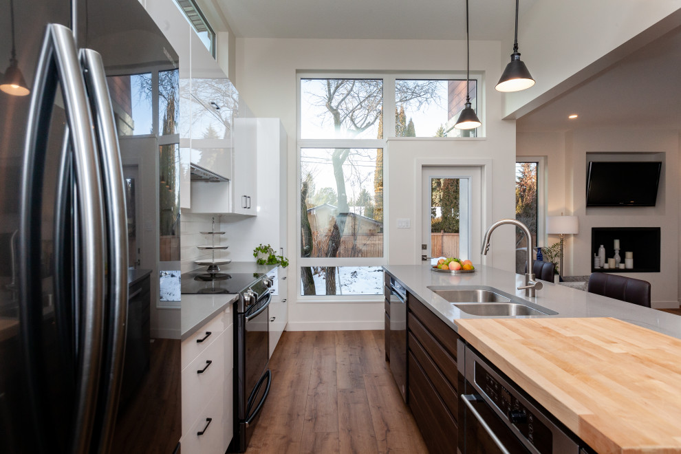 Photo of a mid-sized contemporary galley open plan kitchen in Edmonton with an undermount sink, flat-panel cabinets, white cabinets, solid surface benchtops, white splashback, subway tile splashback, black appliances, vinyl floors, with island, brown floor, grey benchtop and vaulted.