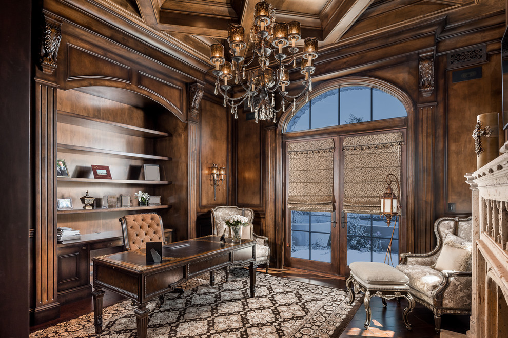 This is an example of an expansive mediterranean home office in Phoenix with a library, beige walls, dark hardwood floors, a standard fireplace, a stone fireplace surround, a freestanding desk, brown floor, coffered and panelled walls.