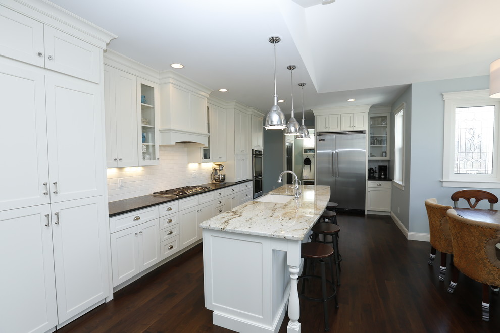 This is an example of a large arts and crafts l-shaped eat-in kitchen in Cedar Rapids with a farmhouse sink, shaker cabinets, white cabinets, granite benchtops, blue splashback, ceramic splashback, stainless steel appliances, dark hardwood floors and with island.