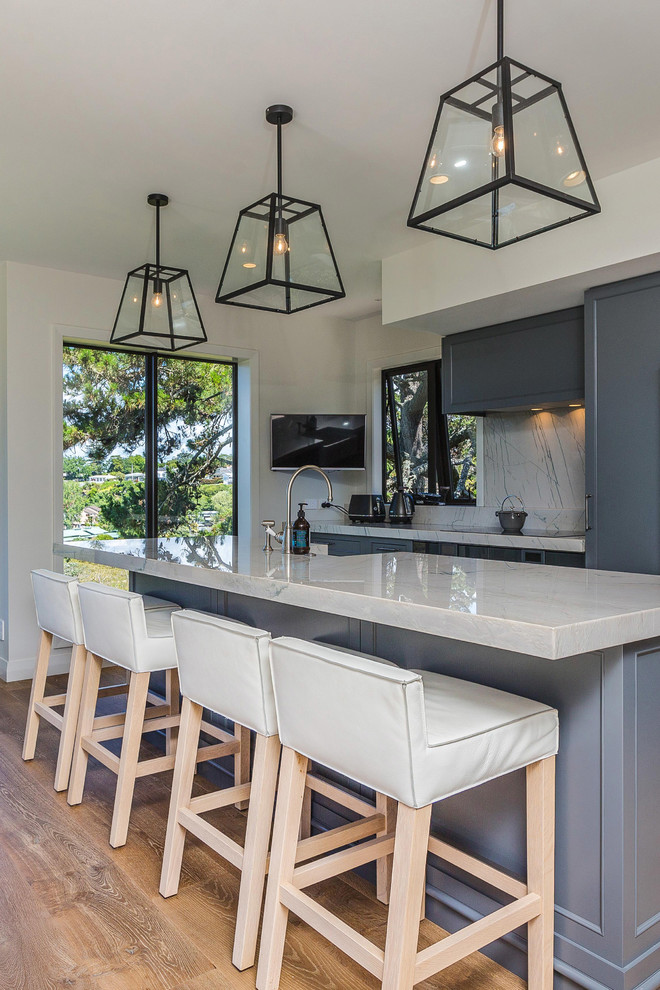Mid-sized transitional galley eat-in kitchen in Auckland with shaker cabinets, blue cabinets, quartzite benchtops, marble splashback, with island, a farmhouse sink, grey splashback, panelled appliances, medium hardwood floors, brown floor and grey benchtop.
