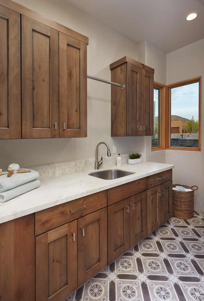 This is an example of a mid-sized l-shaped dedicated laundry room in Phoenix with an undermount sink, recessed-panel cabinets, quartzite benchtops, beige walls, concrete floors, a side-by-side washer and dryer, brown floor, white benchtop and medium wood cabinets.