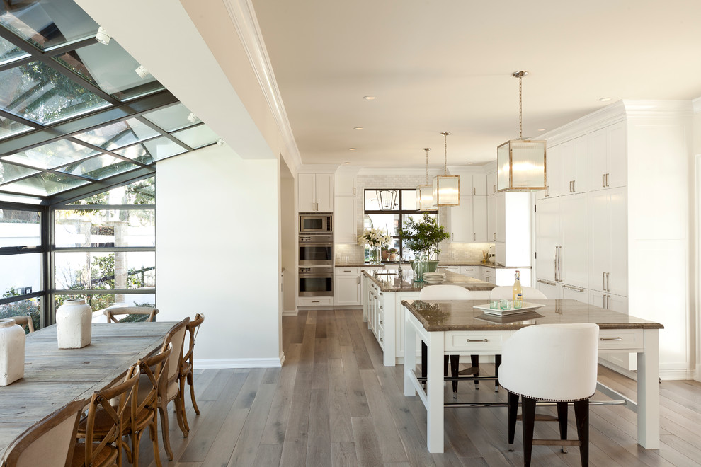 This is an example of a large transitional l-shaped eat-in kitchen in Los Angeles with shaker cabinets, white cabinets, stainless steel appliances, light hardwood floors, multiple islands, a farmhouse sink, marble benchtops, white splashback and glass tile splashback.