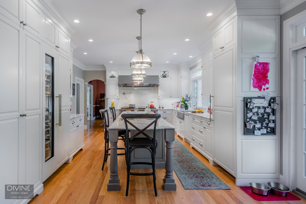 Mid-sized elegant galley light wood floor and brown floor eat-in kitchen photo in Boston with a farmhouse sink, shaker cabinets, white cabinets, marble countertops, white backsplash, subway tile backsplash, stainless steel appliances and an island
