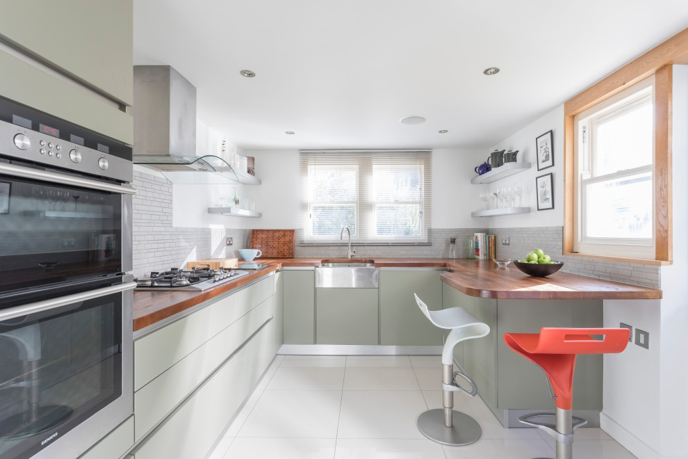 This is an example of a contemporary u-shaped kitchen in Sussex with a farmhouse sink, glass-front cabinets, grey cabinets, wood benchtops, grey splashback, stainless steel appliances, no island, grey floor and brown benchtop.