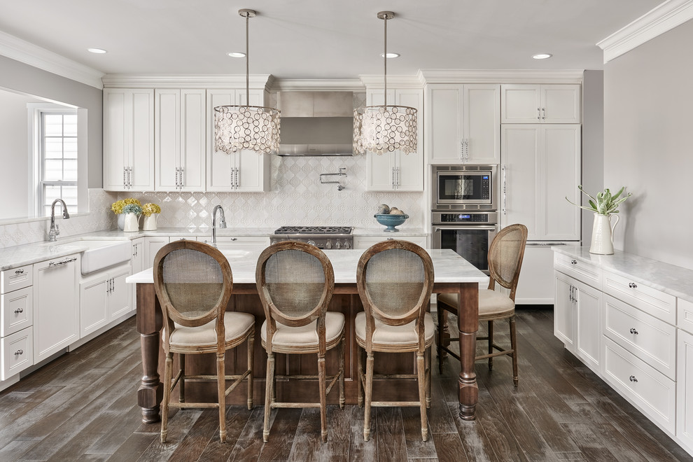 Photo of a large traditional u-shaped open plan kitchen in DC Metro with a farmhouse sink, recessed-panel cabinets, white cabinets, marble benchtops, white splashback, ceramic splashback, stainless steel appliances, medium hardwood floors, with island and grey floor.