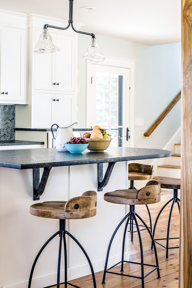 Design ideas for a country l-shaped open plan kitchen in Portland Maine with a farmhouse sink, shaker cabinets, white cabinets, granite benchtops, blue splashback, porcelain splashback, stainless steel appliances, light hardwood floors, with island and black benchtop.
