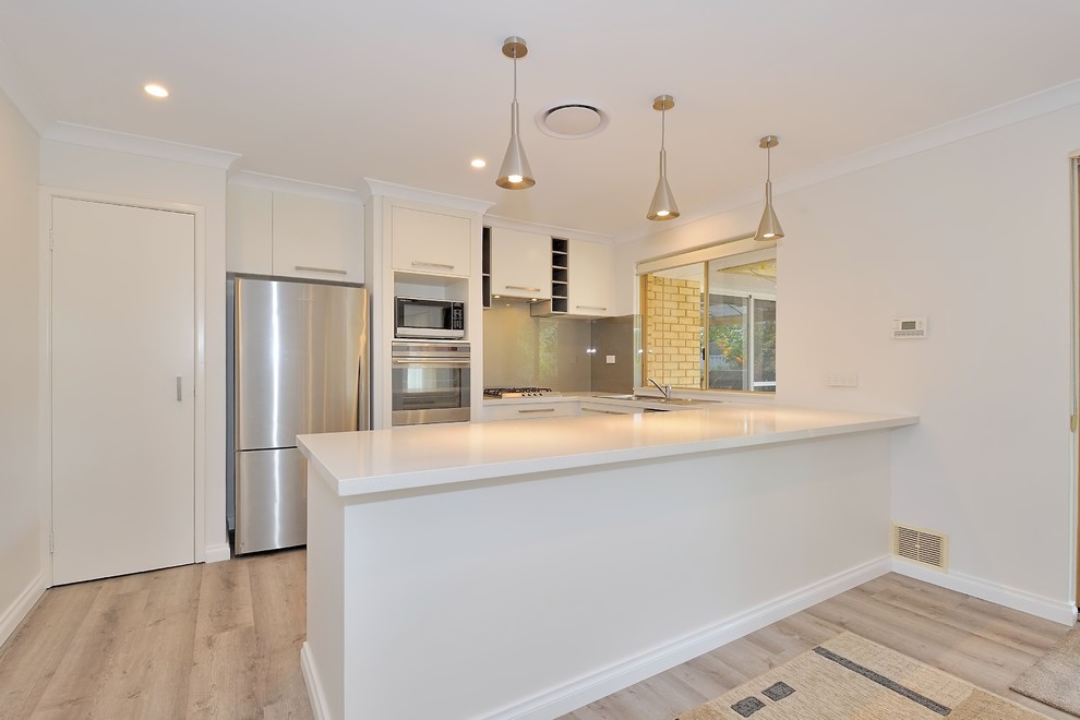 Mid-sized contemporary l-shaped eat-in kitchen in Perth with a double-bowl sink, flat-panel cabinets, white cabinets, quartz benchtops, grey splashback, glass sheet splashback, stainless steel appliances, light hardwood floors, a peninsula, brown floor and white benchtop.