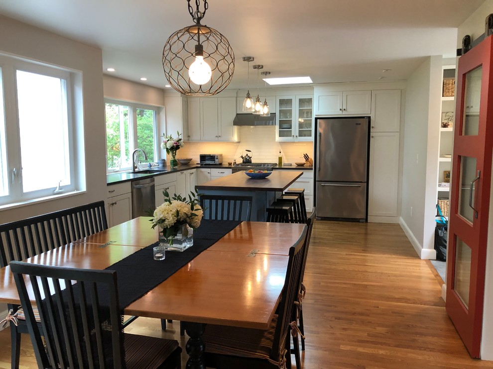 Mid-sized transitional l-shaped medium tone wood floor and brown floor open concept kitchen photo in Boston with a single-bowl sink, shaker cabinets, white cabinets, granite countertops, white backsplash, window backsplash, stainless steel appliances, an island and black countertops