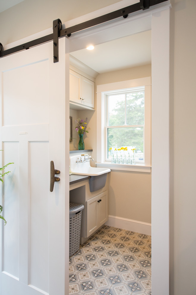 This is an example of a beach style dedicated laundry room in Boston with a farmhouse sink, white cabinets, solid surface benchtops, beige walls, ceramic floors, a side-by-side washer and dryer, multi-coloured floor, grey benchtop and recessed-panel cabinets.