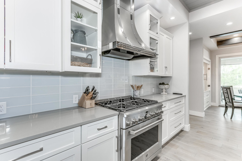 Design ideas for a large modern l-shaped eat-in kitchen in Wichita with a farmhouse sink, shaker cabinets, white cabinets, grey splashback, ceramic splashback, stainless steel appliances, light hardwood floors, with island, brown floor, grey benchtop and granite benchtops.