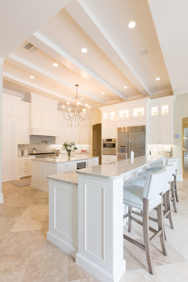 Transitional l-shaped open plan kitchen in Orange County with a single-bowl sink, shaker cabinets, white cabinets, marble benchtops, white splashback, stone slab splashback, stainless steel appliances, travertine floors and multiple islands.