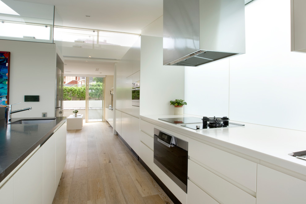 Photo of a contemporary galley kitchen in Sydney with an undermount sink, flat-panel cabinets, white cabinets, window splashback, black appliances, light hardwood floors, with island, beige floor and white benchtop.