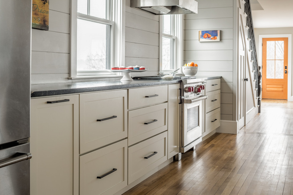This is an example of a mid-sized transitional galley kitchen in Portland Maine with flat-panel cabinets, soapstone benchtops, shiplap splashback, stainless steel appliances, medium hardwood floors and black benchtop.