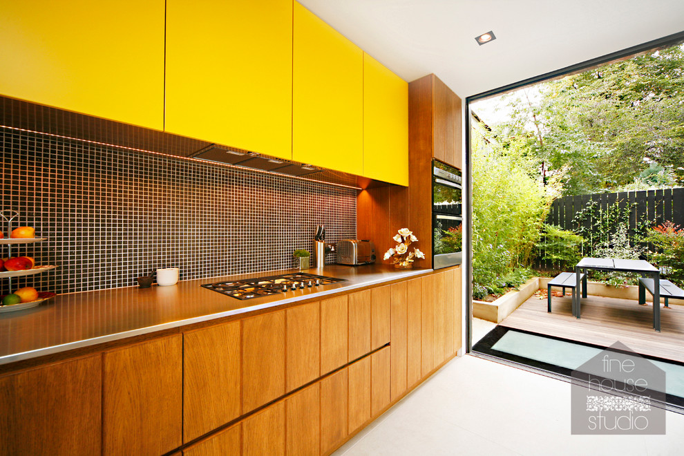 Midcentury single-wall eat-in kitchen in Gloucestershire with yellow cabinets, stainless steel benchtops, black splashback, mosaic tile splashback, stainless steel appliances and porcelain floors.