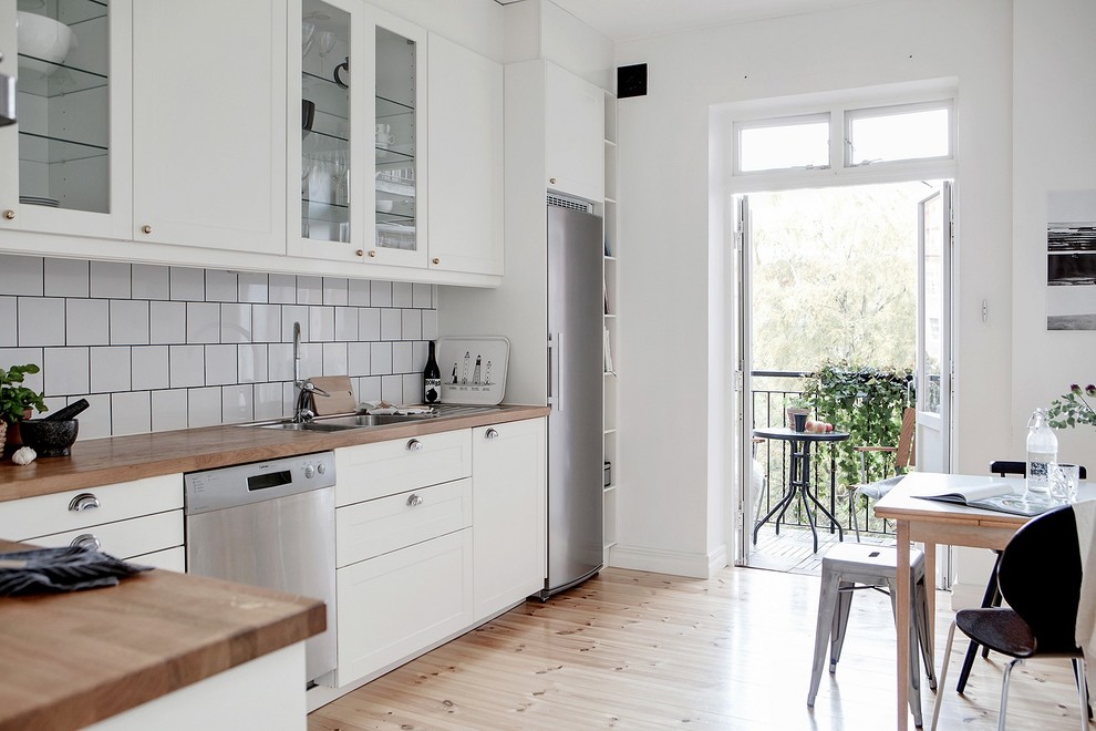 Scandinavian l-shaped eat-in kitchen in Malmo with a double-bowl sink, shaker cabinets, white cabinets, wood benchtops, white splashback, stainless steel appliances, light hardwood floors and beige floor.
