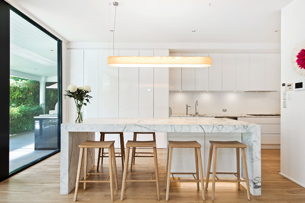 This is an example of a contemporary open plan kitchen in Adelaide with white cabinets, marble benchtops, white splashback, stainless steel appliances, light hardwood floors and with island.