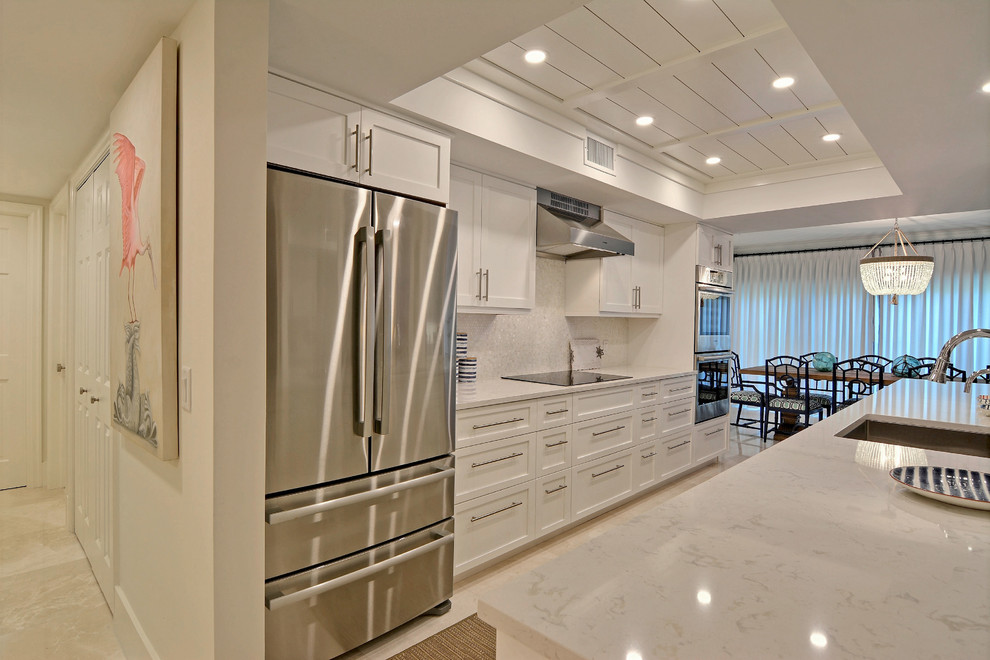 Photo of a small beach style galley eat-in kitchen in Miami with a single-bowl sink, recessed-panel cabinets, white cabinets, white splashback, porcelain splashback, stainless steel appliances, with island and white benchtop.
