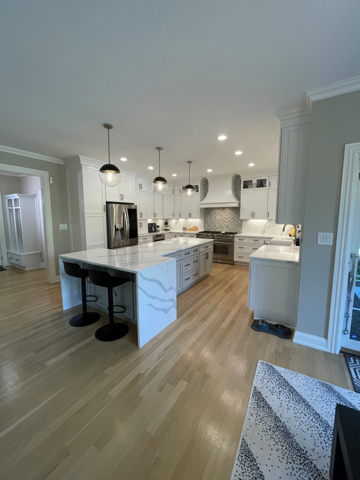 Example of a large trendy u-shaped light wood floor and beige floor eat-in kitchen design with a farmhouse sink, shaker cabinets, white cabinets, quartz countertops, gray backsplash, quartz backsplash, stainless steel appliances, an island and white countertops