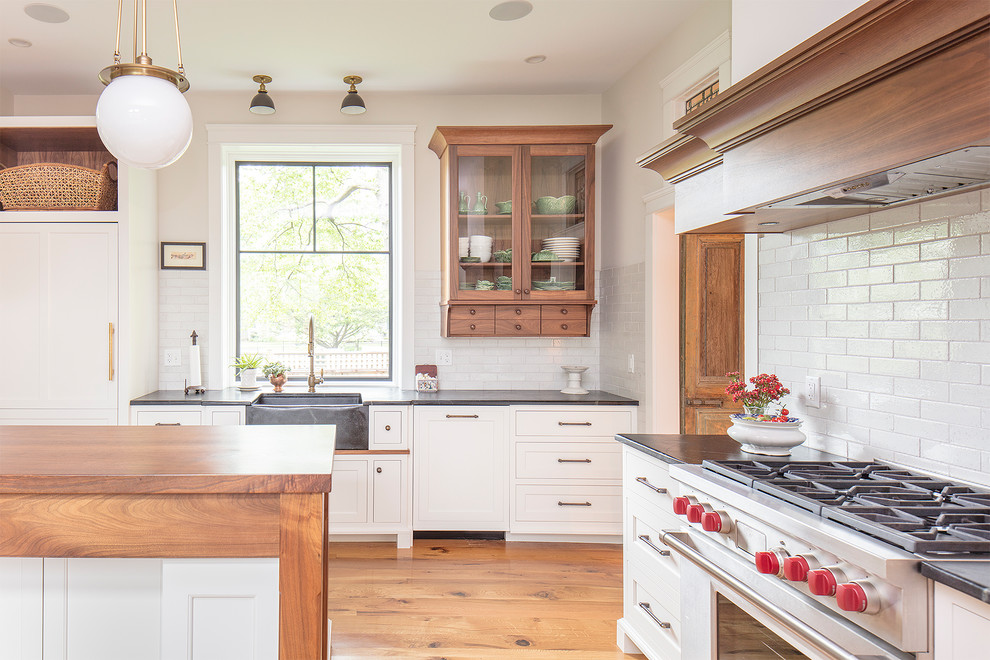 This is an example of a mid-sized country l-shaped kitchen in Philadelphia with a farmhouse sink, shaker cabinets, white cabinets, soapstone benchtops, white splashback, brick splashback, panelled appliances, medium hardwood floors, with island, brown floor and black benchtop.