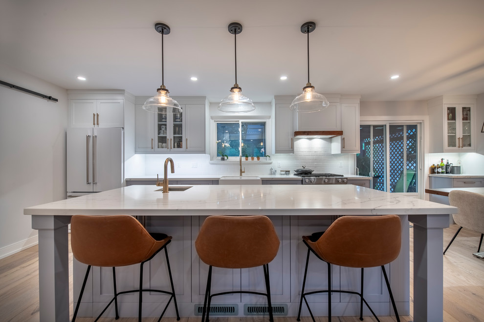 Inspiration for a contemporary galley eat-in kitchen in Toronto with a farmhouse sink, shaker cabinets, white cabinets, white splashback, brick splashback, light hardwood floors, with island, beige floor, white benchtop, quartz benchtops and stainless steel appliances.