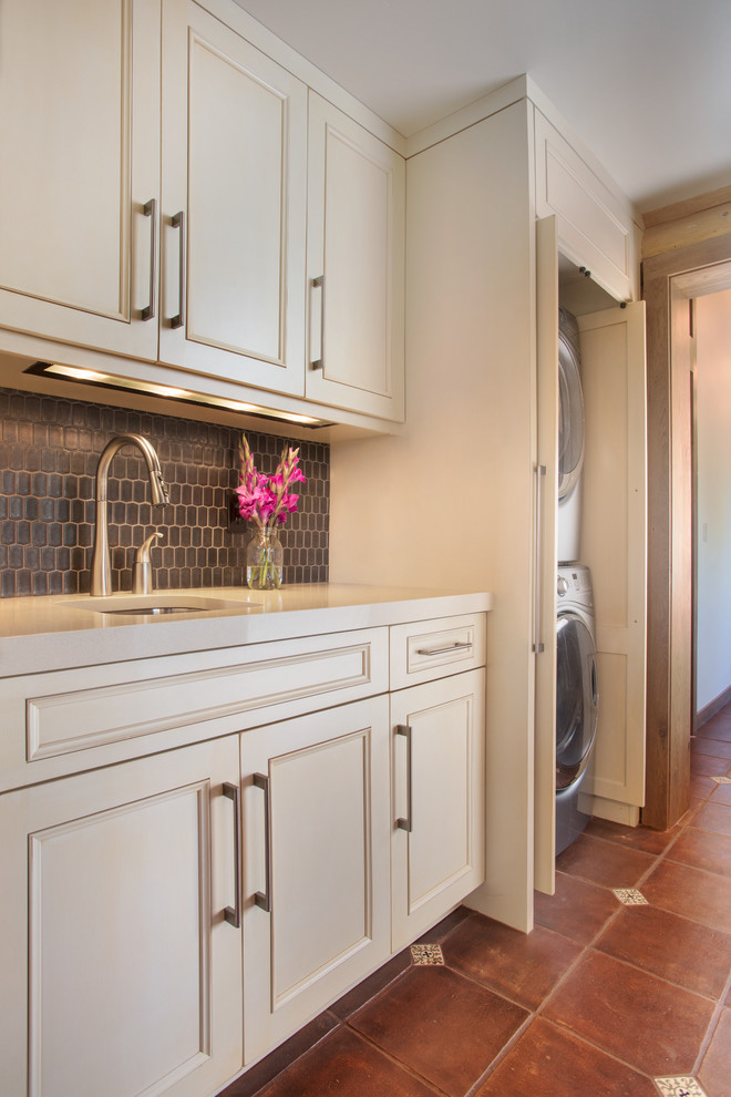 This is an example of a mid-sized transitional single-wall laundry room in Denver with an undermount sink, recessed-panel cabinets, white cabinets, solid surface benchtops, beige walls, ceramic floors, a stacked washer and dryer, brown floor and beige benchtop.