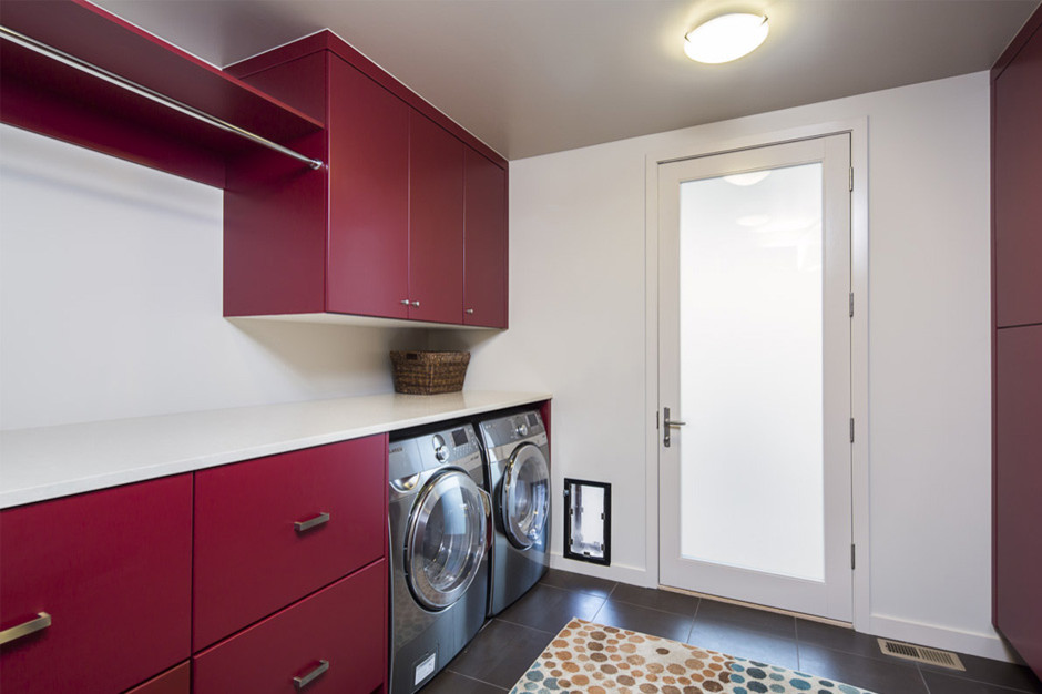 Photo of a mid-sized modern galley dedicated laundry room in Denver with flat-panel cabinets, red cabinets, white walls, a side-by-side washer and dryer, solid surface benchtops, porcelain floors and black floor.
