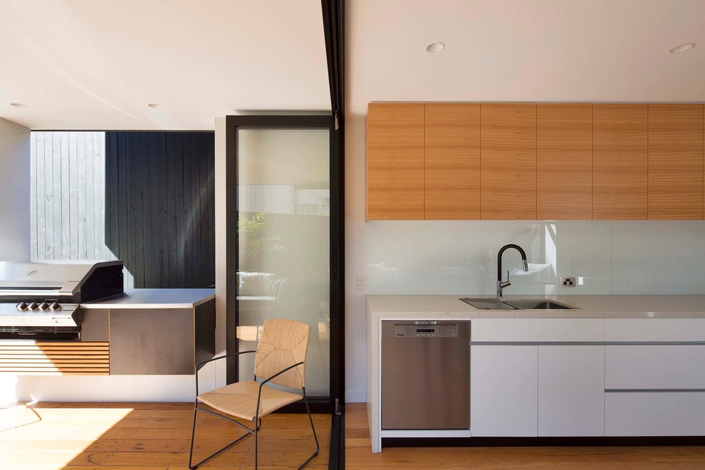 This is an example of a modern single-wall eat-in kitchen in Sydney with an undermount sink, flat-panel cabinets, quartz benchtops, white splashback, glass sheet splashback, black appliances, medium hardwood floors and no island.