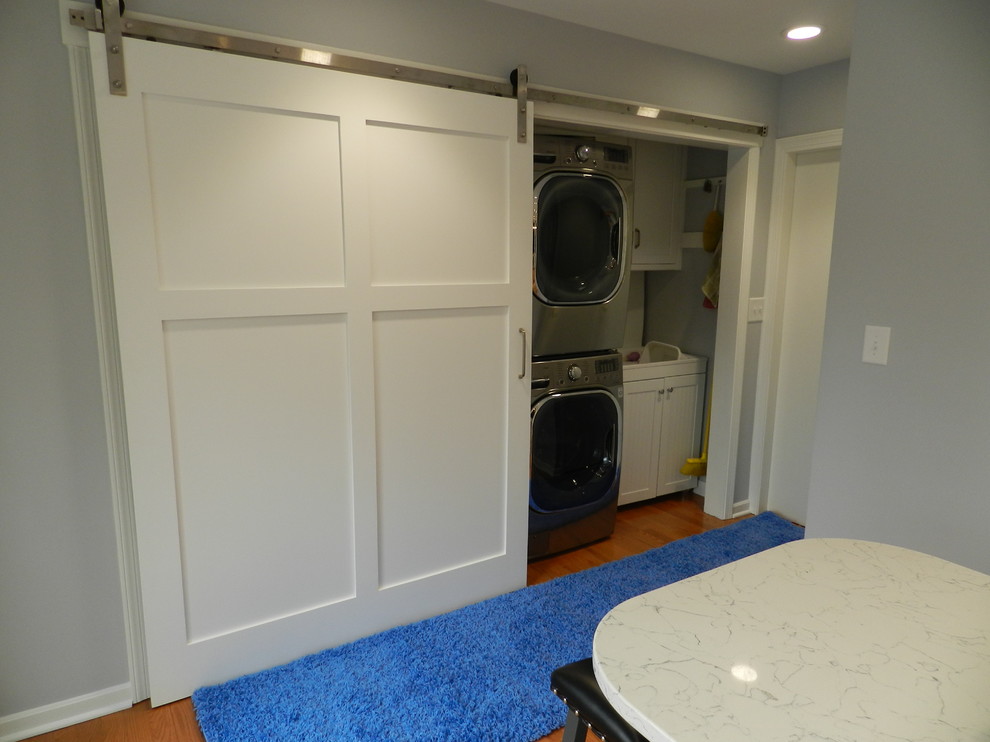 Mid-sized country single-wall laundry cupboard in Cleveland with an integrated sink, recessed-panel cabinets, white cabinets, solid surface benchtops, beige walls, light hardwood floors, a stacked washer and dryer and beige floor.
